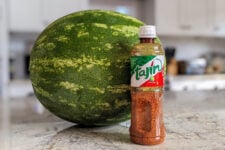 A bottle of Tajin chili lime seasoning with a watermelon behind it.