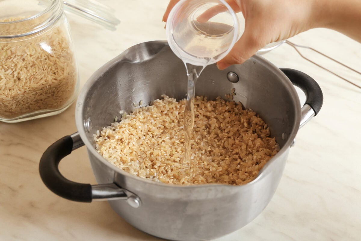 A woman's hand pouring water into a pot of uncooked brown rice with a jar of brown rice in the background.