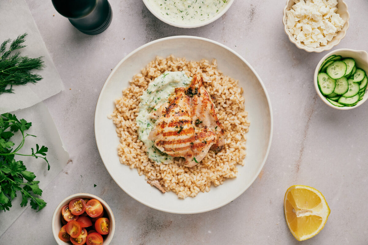 Assembling a Greek Chicken Bowl by layering brown rice, Tzatziki, and chicken.