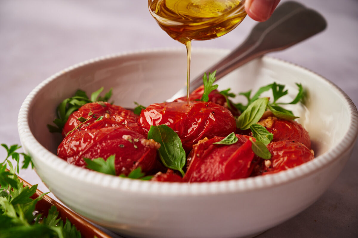 Oven roasted tomatoes in a white bowl garnished with basil with a hand drizzling olive oil on top.