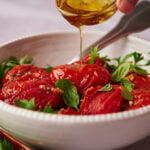 Oven roasted tomatoes in a white bowl garnished with basil with a hand drizzling olive oil on top.