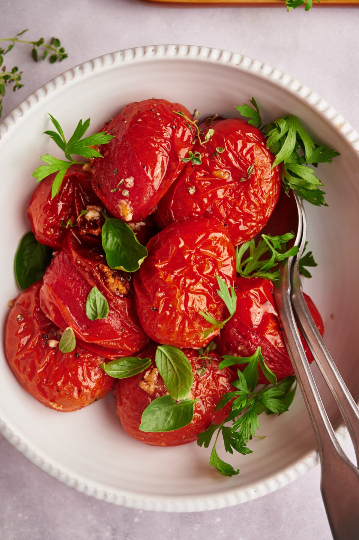 Oven roasted tomatoes in a white bowl garnished with basil and two spoons.
