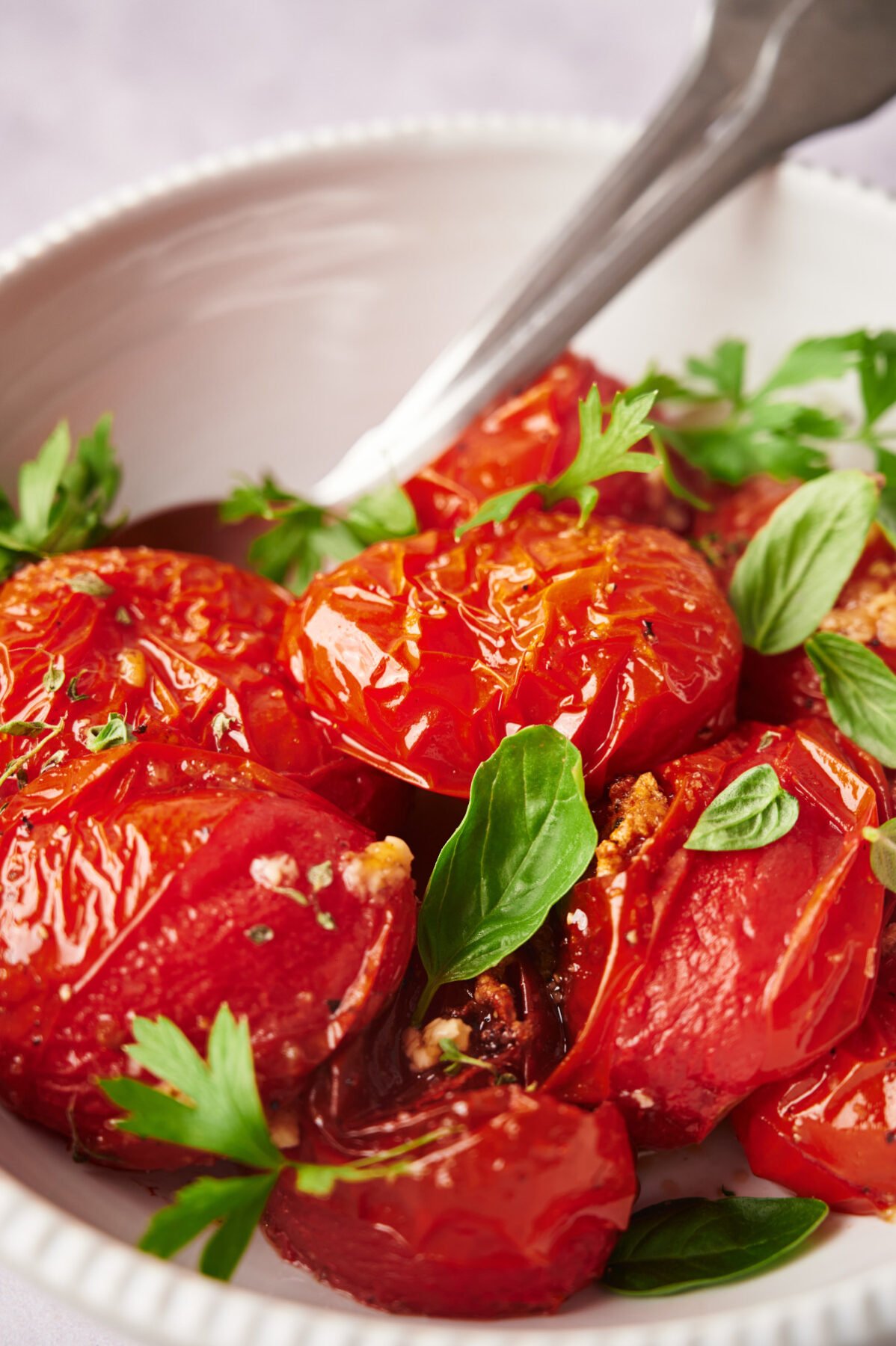 Oven roasted tomatoes in a white bowl garnished with basil and two spoons.