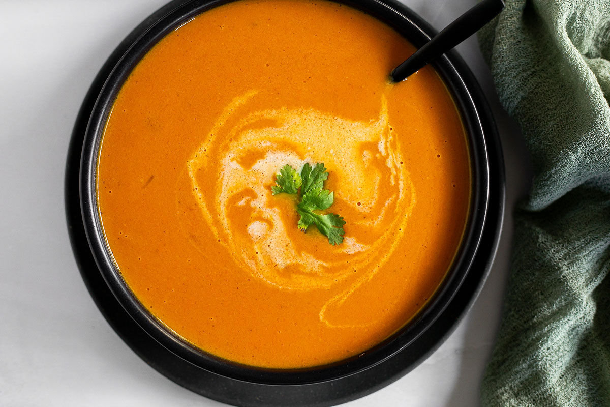 A black bowl filled with coconut carrot soup, garnished with fresh cilantro, with a spoon and a green napkin. 