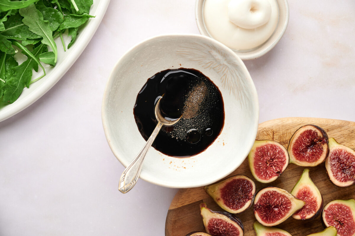 Balsamic vinegar with maple syrup, salt, and pepper being mixed in a bowl with sliced figs in the background.