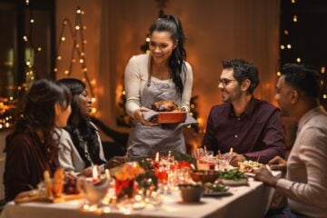 People at a table with holiday decor and a woman holding a dish in her hands ready to serve it to her guests.