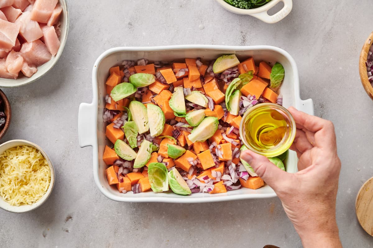 A woman's hand drizzling olive oil over a casserole dish filled with fall harvest vegetables.