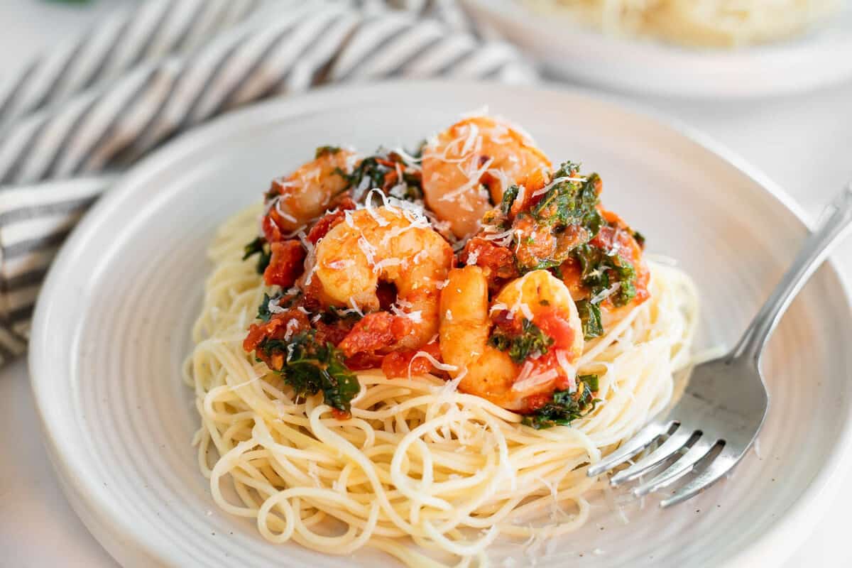 Spicy shrimp and kale served over angel hair pasta on a white plate with a fork and a striped napkin.