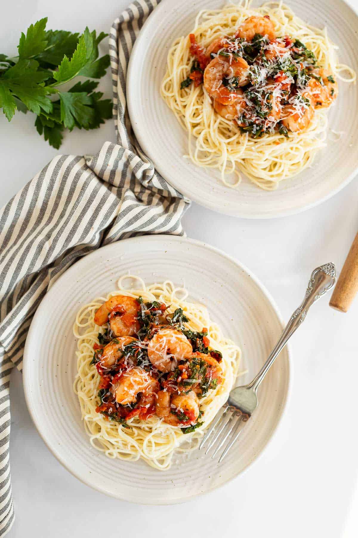 Spicy shrimp and kale served over angel hair pasta on white plates with a fork and a stripped napkin.