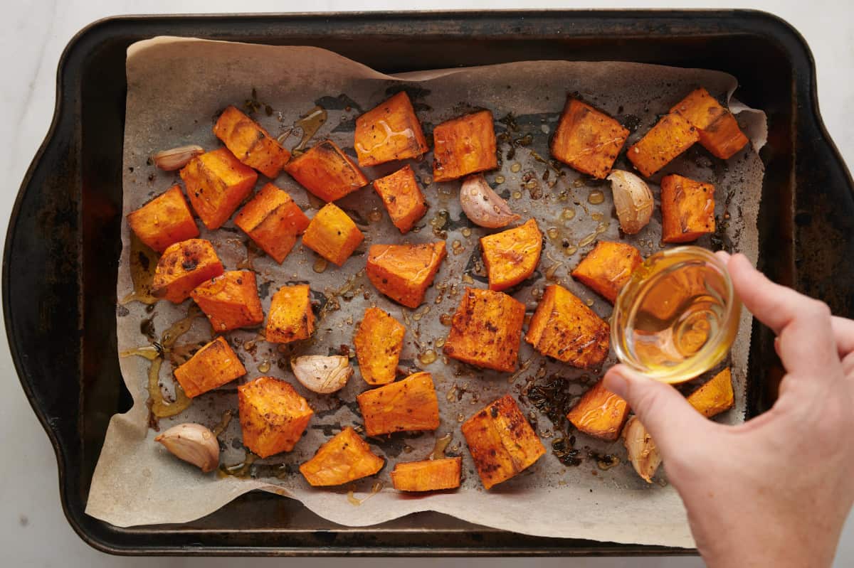 Pouring honey over sweet potatoes and garlic as they cook on a parchment lined baking sheet.