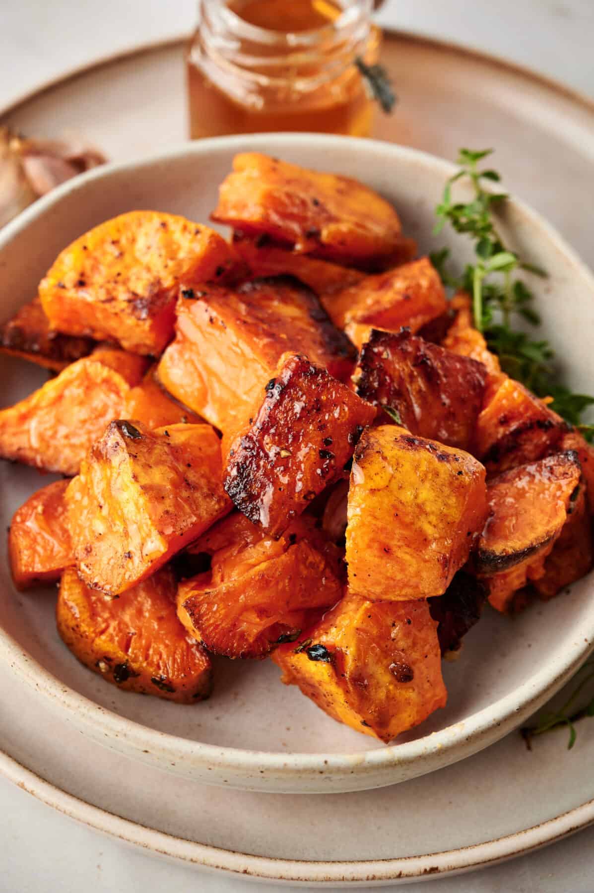 Vertical photo of Cinnamon Roasted Sweet Potatoes in a white dish garnished with thyme with honey in the background.