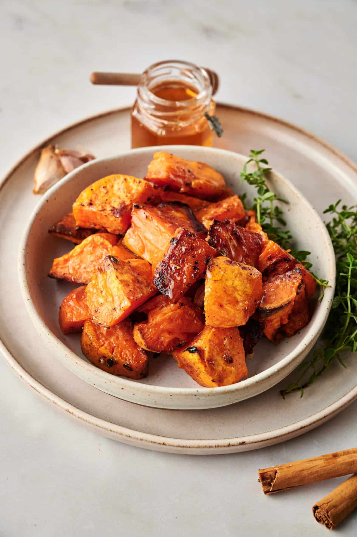 Cinnamon Roasted Sweet Potatoes in a white dish garnished with thyme with honey in the background.
