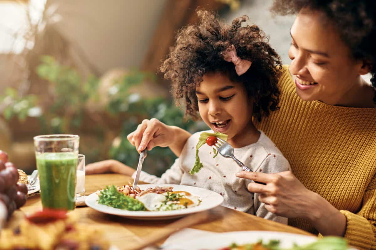 Happy African American girl sitting on mother's lap while she is feeding her healthy seasonal vegetables at dining table.