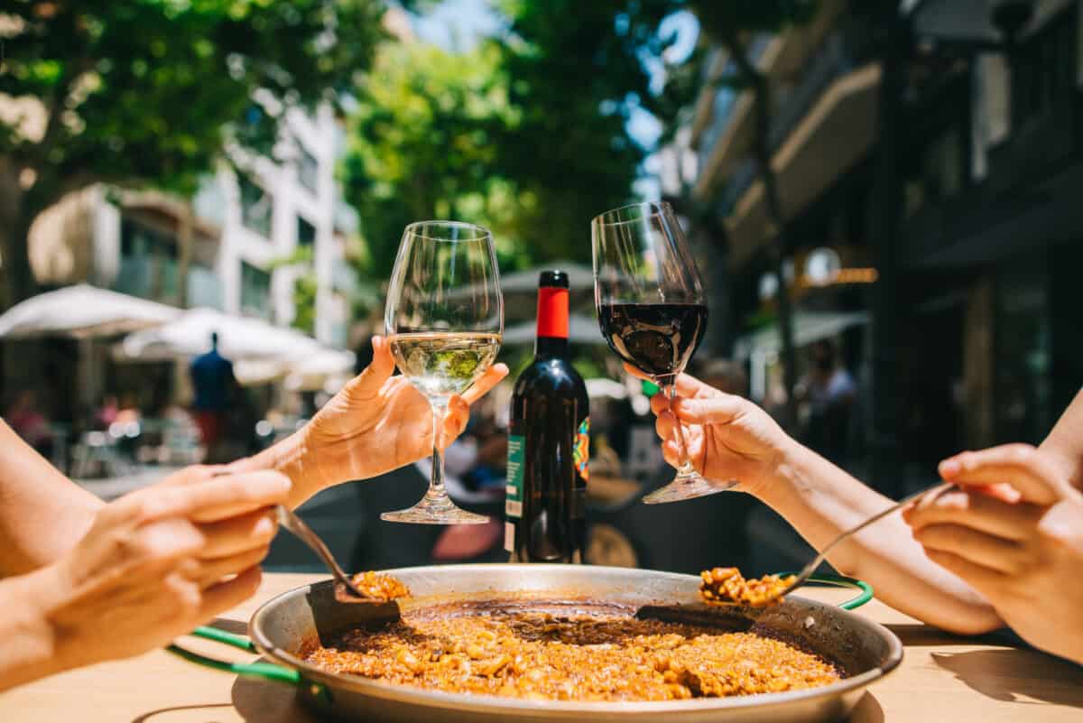 Two women holding drinks wine start eating paella with seafood and squid at a table in a Mediterranean restaurant.