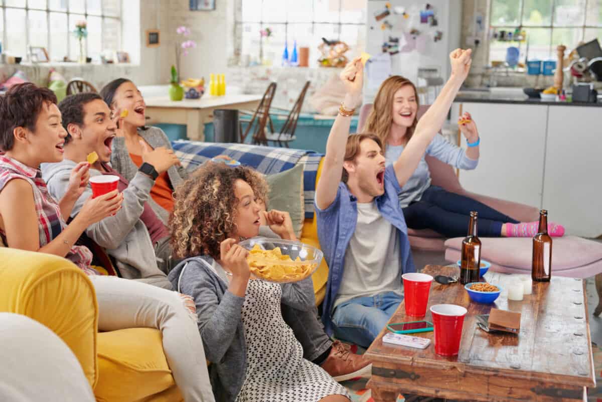 Diverse mix of friends sports fans watching football game on TV at home, celebrating winning touchdown huddled on couch shouting excited sharing snacks drinking beer.