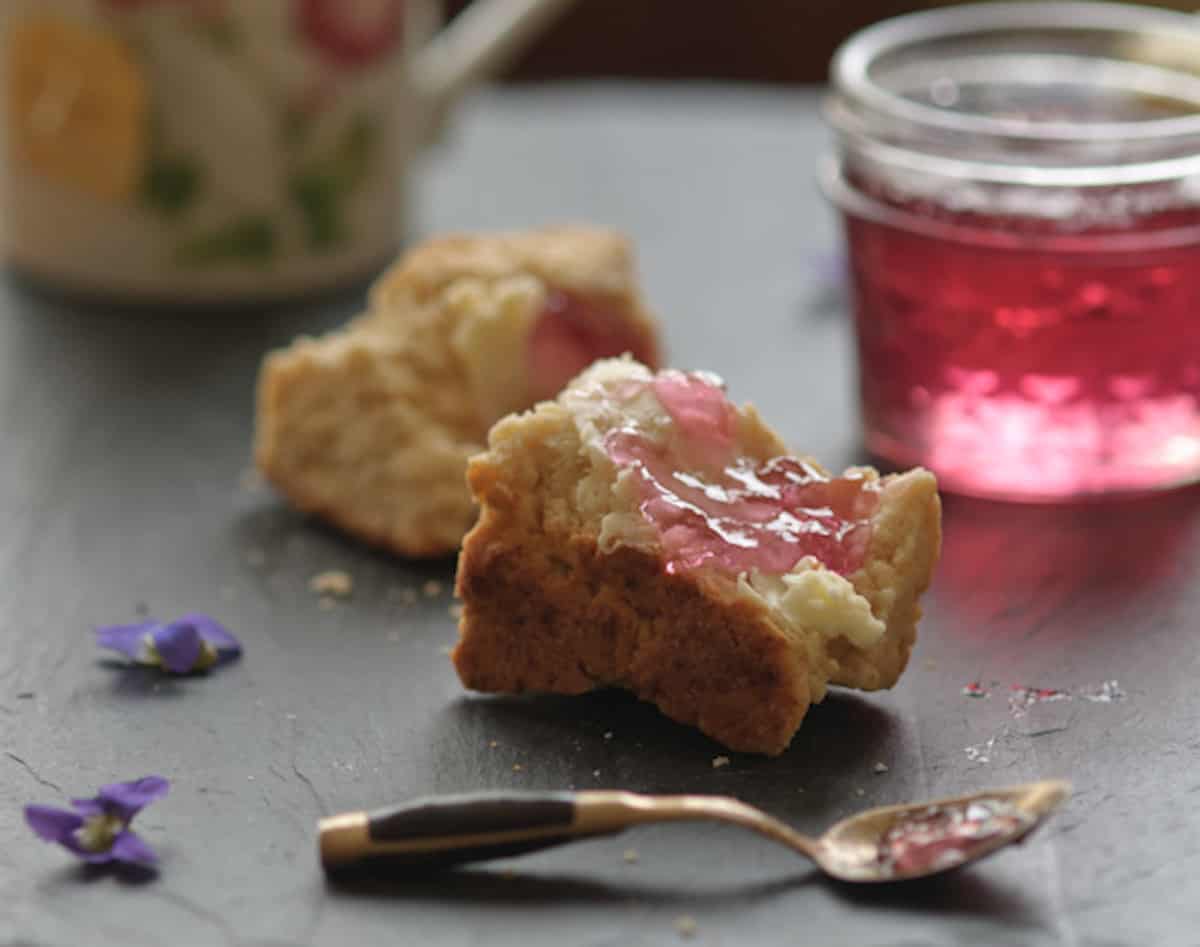 Violet jelly spread on a biscuit with a spoon in front and a jar of violet jelly in the background.