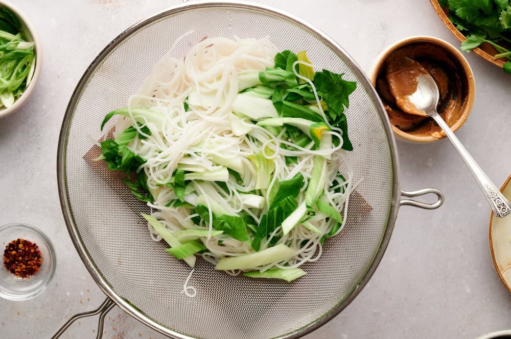 Rice noodles being mixed with blanched Bok Choy in a strainer.
