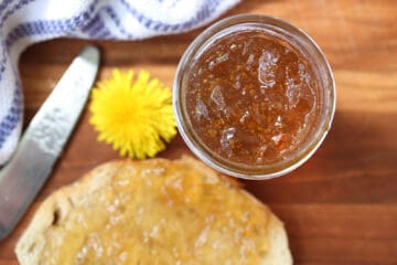 A close-up view of an open jar of dandelion jelly with a dandelion flower and jelly spread on toast in the background.